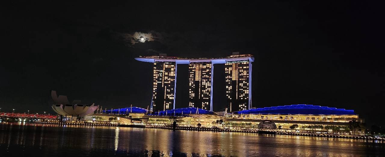 Marina Bay Sands and the ArtScience Museum at night, lit blue and gold, reflected in the water under a moon.