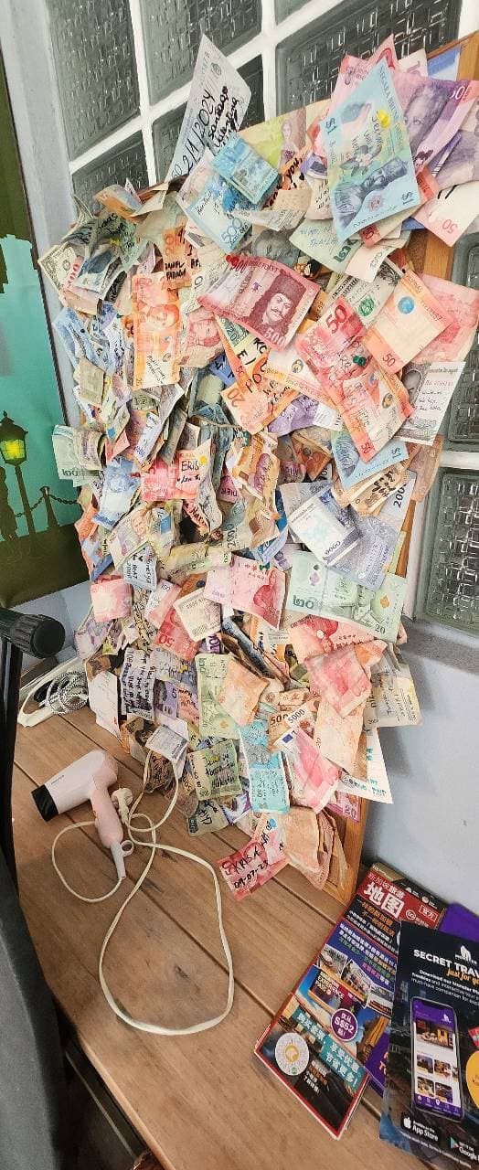 A hostel wall at Boat Quay covered in banknotes from other countries, with a hairdryer on the table below. 16/02/2025 Nacnano was here.